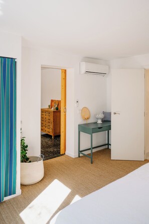 Une pièce comprenant un lit, un bureau avec une chaise, une commode, un appareil de climatisation et une porte.