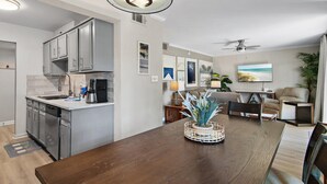 Een moderne keuken met grijze kastjes, een ingebouwde vaatwasser en een koffiezetapparaat. Er is een eethoek met een houten tafel en stoelen, een plant als middelpunt en een plafondventilator.
