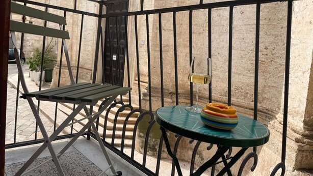 A balcony with a green folding table, a small round table with a colorful stack of plates, and a glass of white wine. - Amazing 2-bedroom apartment in adorable Monopoli with AC (Monopoli)