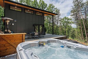 Ein moderner Außenbereich mit Whirlpool, einer Holzterrasse, einer überdachten Lounge und Blick auf eine bewaldete Landschaft.