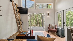Ein modernes Wohnzimmer mit einer Couch, einem Couchtisch und einem an der Wand befestigten Fernseher. Eine Leiter lehnt an der Wand, und eine Tür führt auf einen Balkon mit Blick auf Bäume.