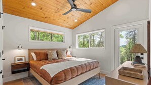 Ein Schlafzimmer mit Holzbalkendecke, einem Bett mit brauner Bettwäsche, zwei Nachttischen, einer Nachttischlampe und Blick auf Bäume durch Fenster.