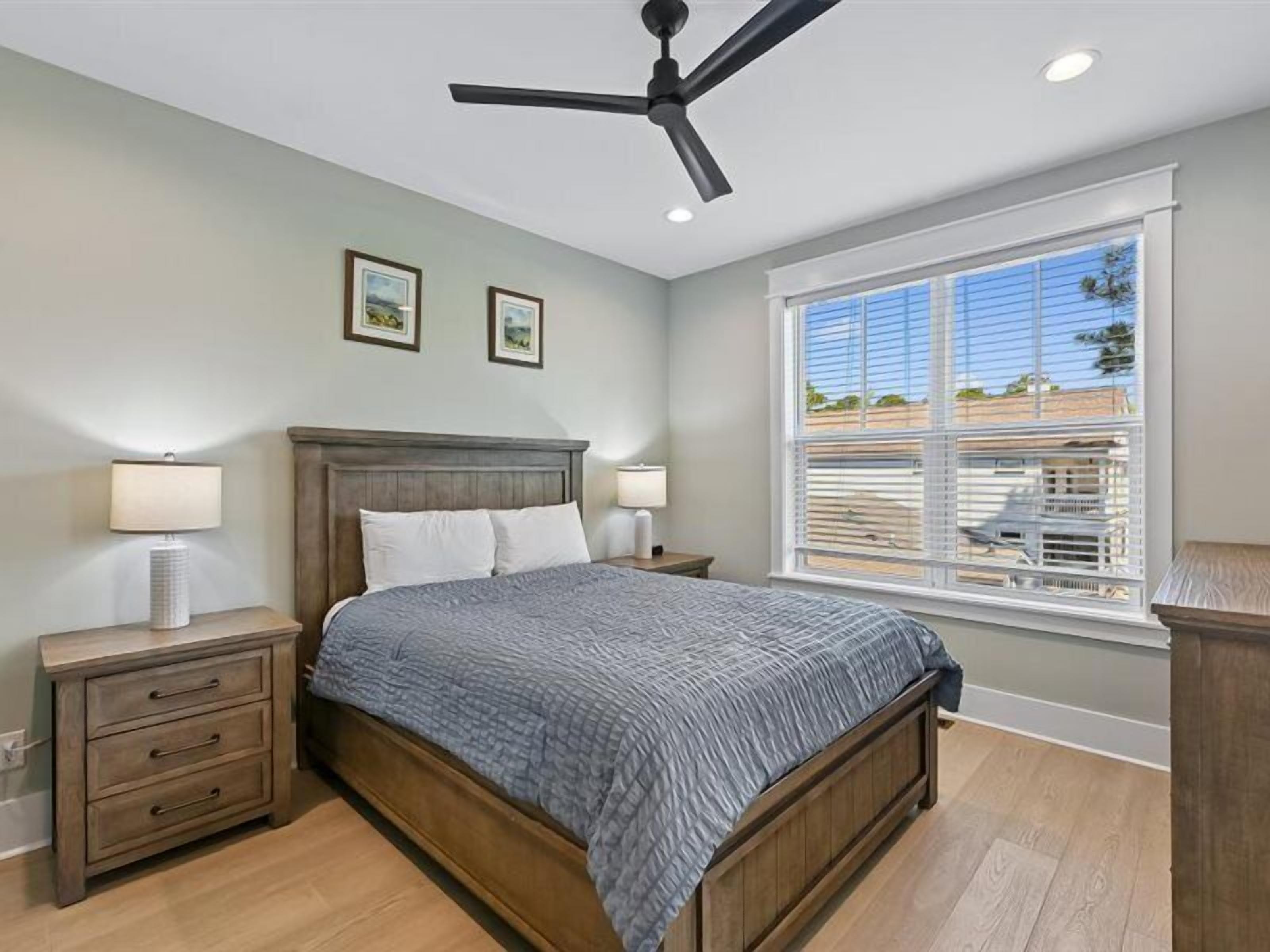 Une chambre à coucher comprenant un lit, des tables de chevet, un ventilateur de plafond, une fenêtre avec des stores et des tableaux encadrés accrochés au mur.