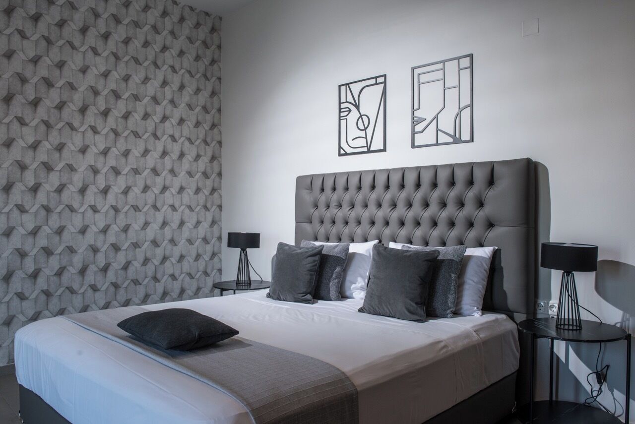 A modern bedroom with a tufted headboard, a neatly made bed with multiple pillows, a patterned wall, and two wall-mounted abstract art pieces.