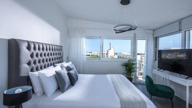 A modern hotel room with a large bed, a TV, a desk, and a view of the cityscape. - Loggia Suites (Heraklion)