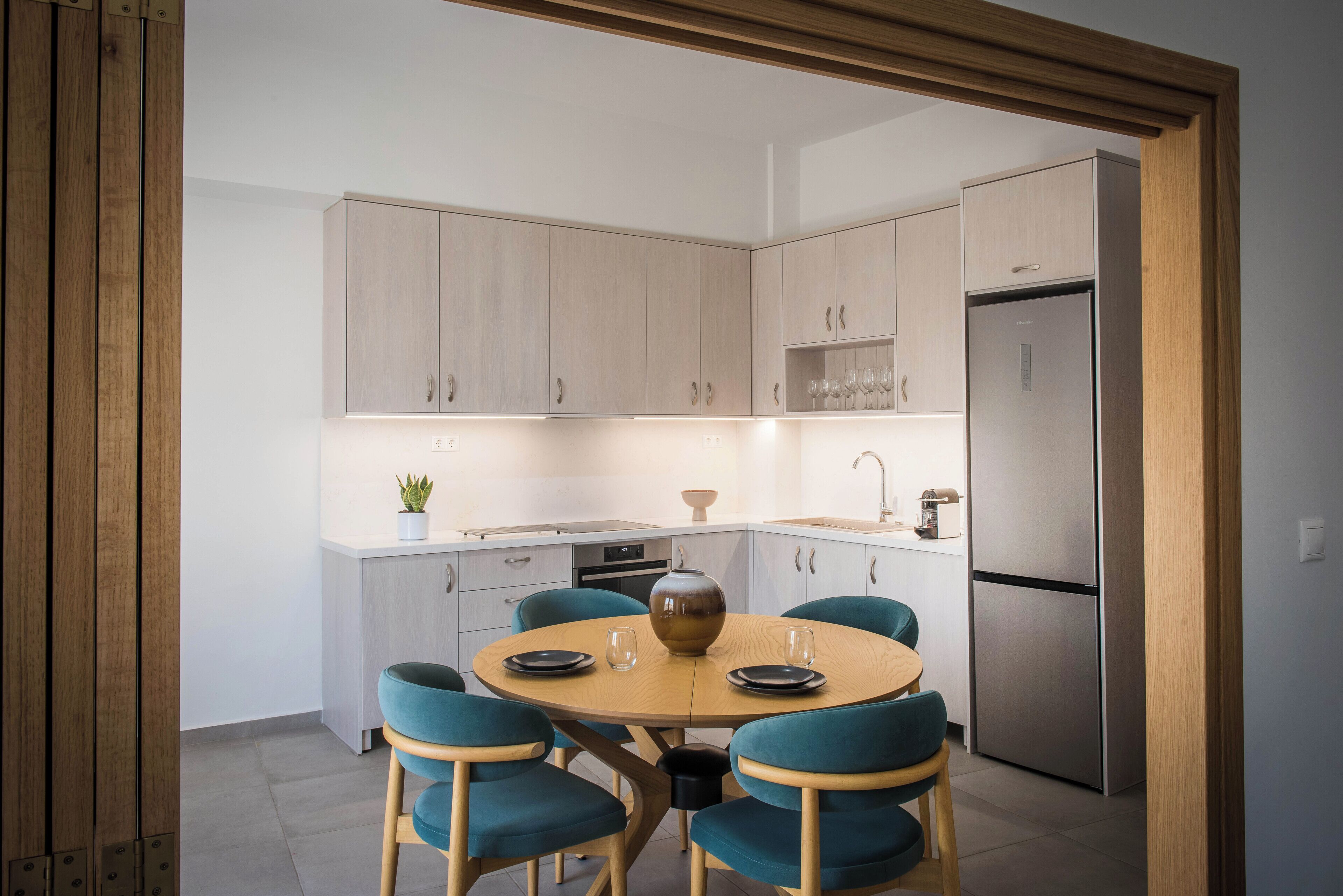 A modern kitchen with light wood cabinets, a stainless steel refrigerator, and teal chairs around a wooden table.