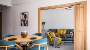 A modern living room with a round wooden table, teal chairs, a sofa with yellow and gray cushions, and a painting on the wall. - Loggia Suites (Heraklion)