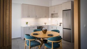 A modern kitchen with light wood cabinets, a stainless steel refrigerator, and a round table with blue chairs. - Loggia Suites (Heraklion)