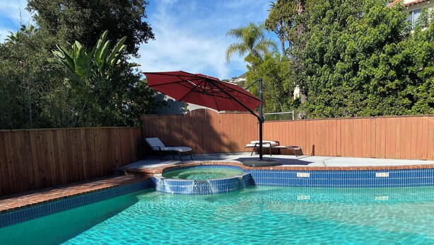 Una piscina con un jacuzzi, rodeada por una valla de madera, y una sombrilla roja que proporciona sombra.