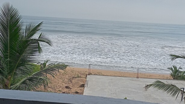 A beach view from a balcony with a wooden ceiling, palm trees, and a sandy beach.