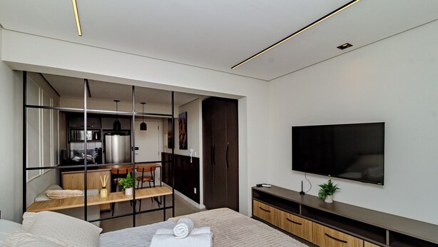 A modern bedroom with a bed, a TV, a desk, and a kitchenette. - Loft Moderno Centro Itajaí (Itajaí)