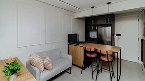 A compact living space with a kitchenette, a grey sofa with decorative pillows, a wooden dining table with two bar stools, and a potted plant on a side table. - Loft Moderno Centro Itajaí (Itajaí)