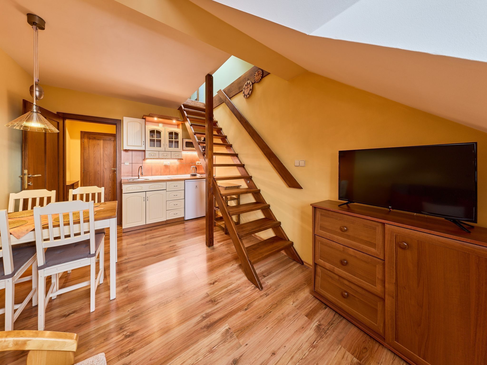 Un vaste espace de vie agrémenté d’un escalier en bois menant à un niveau supérieur, un coin repas avec des chaises blanches et un téléviseur à écran plat posé sur un meuble en bois.
