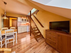 Ein großzügiger Wohnbereich mit einer Holztreppe zu einer oberen Ebene, einem Essbereich mit weißen Stühlen und einem Flachbildfernseher auf einem Holzkasten.