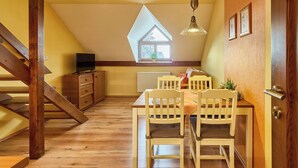 A cozy attic room with a sloped ceiling, wooden staircase, a dining table with chairs, a television on a cabinet, and a window with a view of greenery. - Vysoké nad Jizerou by Interhome (Vysoké nad Jizerou)