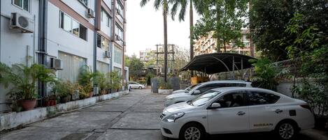 A parking area with two white cars, a covered structure, and potted plants along the building's facade.