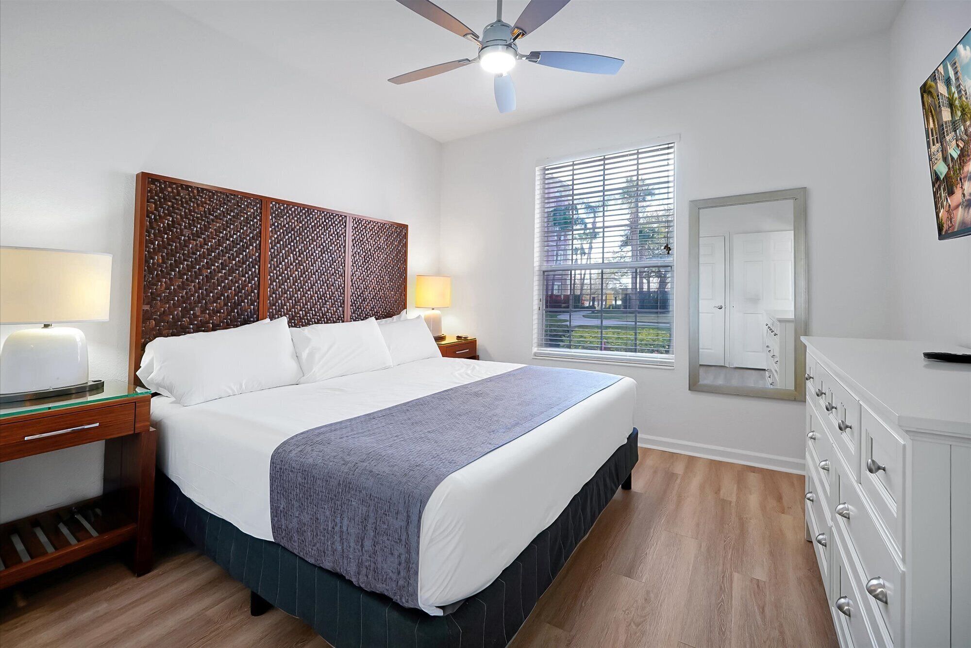 A hotel room with a bed, bedside table, lamp, ceiling fan, window with blinds, dresser, and a mirror.