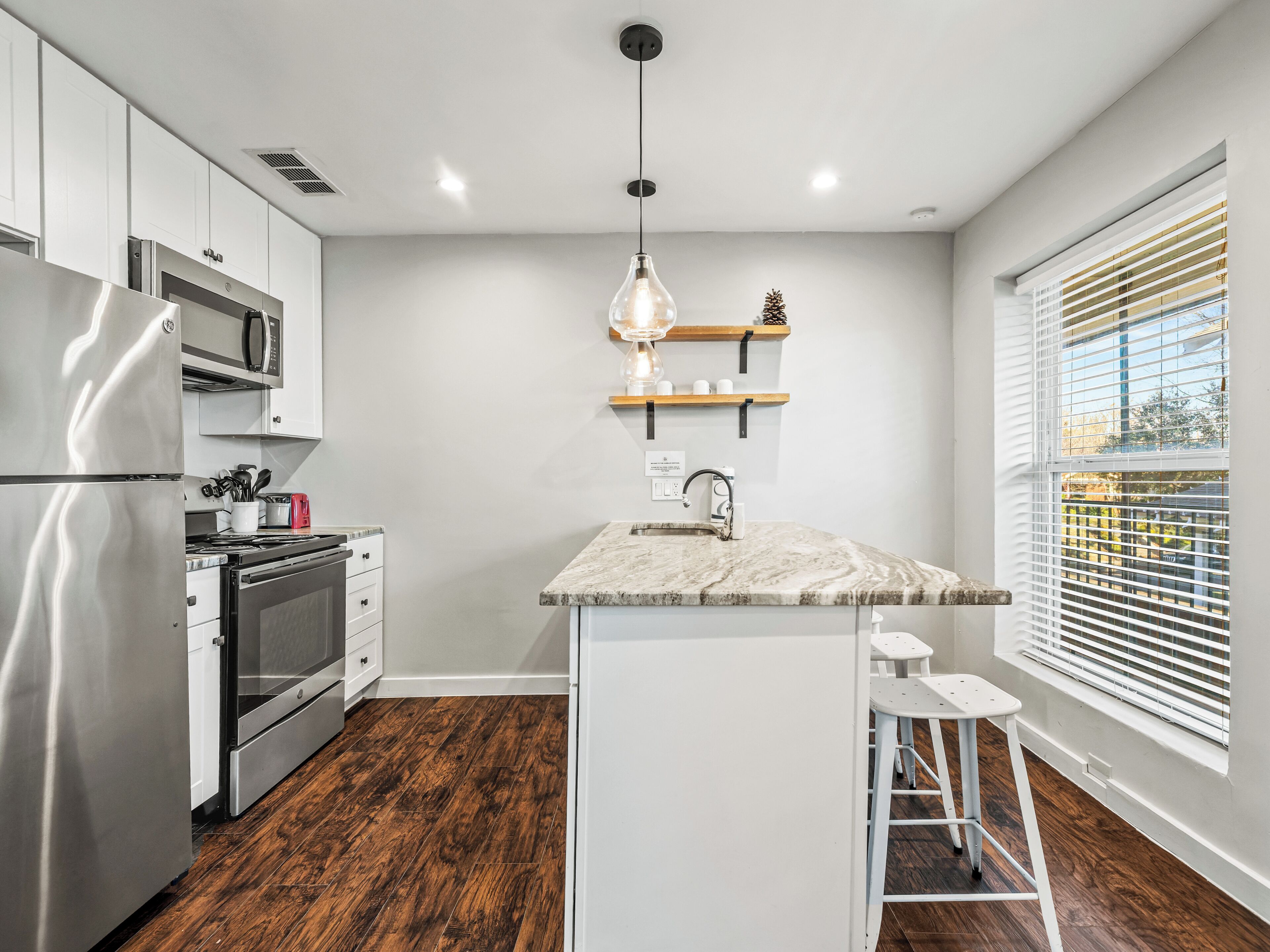 Una cocina moderna con armarios blancos, electrodomésticos de acero inoxidable, piso de madera, una isla central con taburetes de bar y una ventana con persianas.