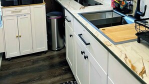 Una cocina moderna con armarios blancos, salpicadero de mármol, microondas negro, cafetera y un lavamanos con tabla de cortar.