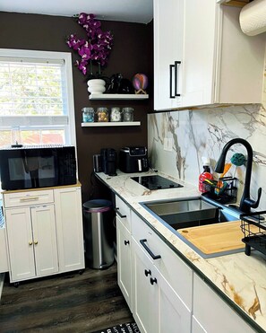 A modern kitchen with white cabinets, a marble backsplash, a black microwave, a coffee maker, and a sink with a cutting board. - Cozy Private Entry Studio • Kitchenette • Security Cam• 8 Min to Stone Mtn  Park (Lilburn)