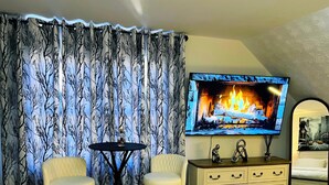 Una sala de estar con una televisión que muestra una chimenea, dos sillones, una mesita y un aparador con cajones.