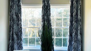 Una habitación con dos sillas blancas, una planta en maceta y una ventana con cortinas estampadas.