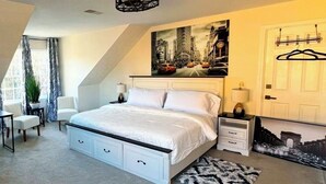 Un dormitorio con una cama grande, un ventilador de techo, un cuadro de un paisaje urbano, una puerta con un perchero y una mesita con una lámpara.