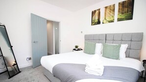 Un dormitorio con una cama grande, cabecera gris, ropa de cama blanca y almohadas estampadas de color verde. Hay una mesita de noche con una planta y un mueble auxiliar con una lámpara. Una puerta conduce a otra habitación.