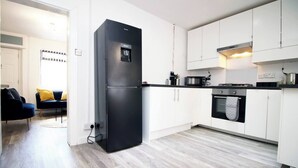 Una cocina moderna con armarios blancos, un refrigerador negro y un microondas. Hay una zona de comedor con una silla azul y un cojín amarillo.