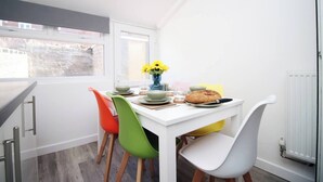 Un pequeño comedor con una mesa blanca, sillas de colores y una ventana con vistas a edificios.