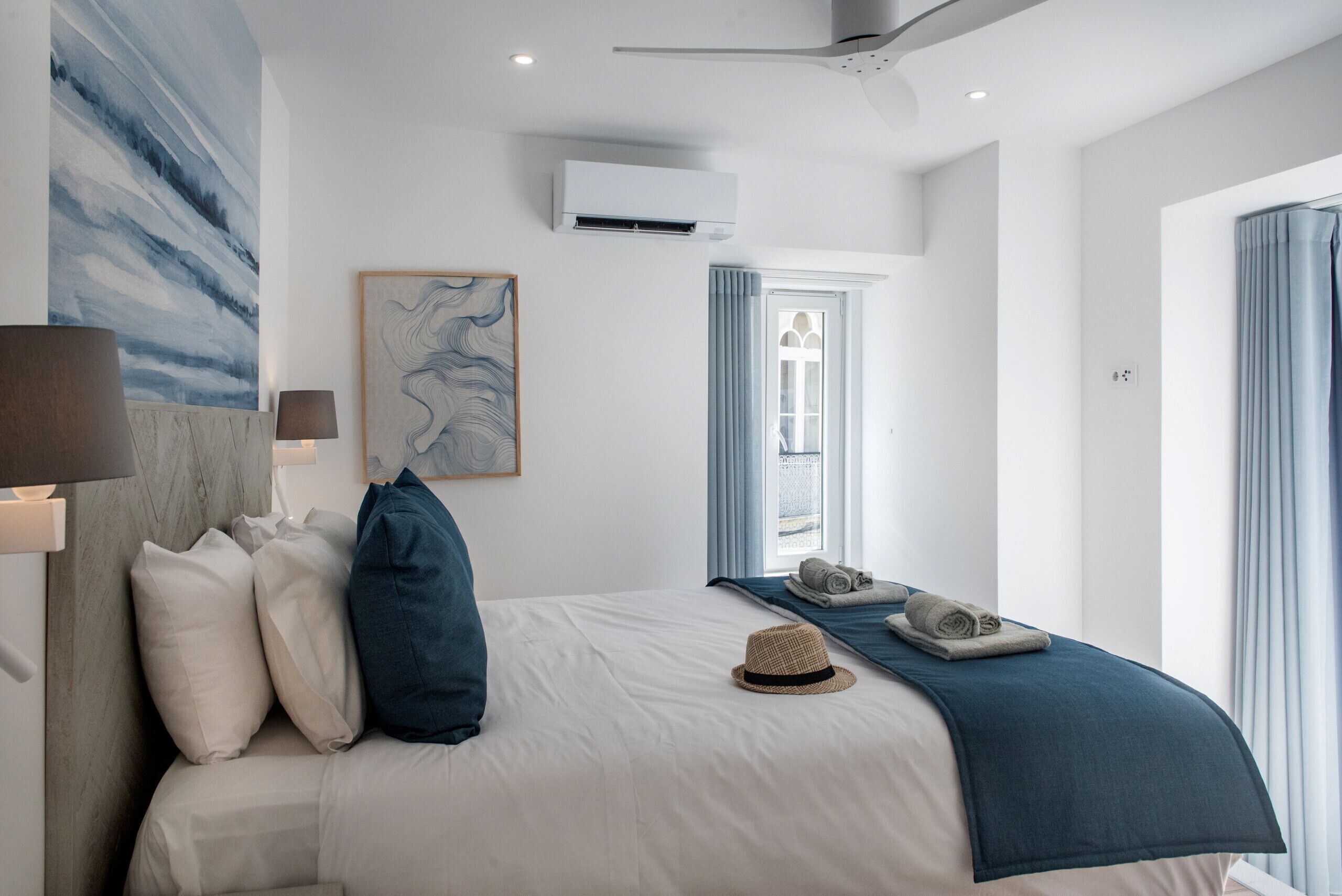A modern hotel room with a bed, a wall-mounted air conditioner, a window with curtains, and a painting.