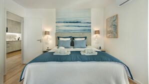 Quarto de hotel moderno com uma cama grande, duas luminárias de cabeceira, um quadro na parede e vista para a área da cozinha.
