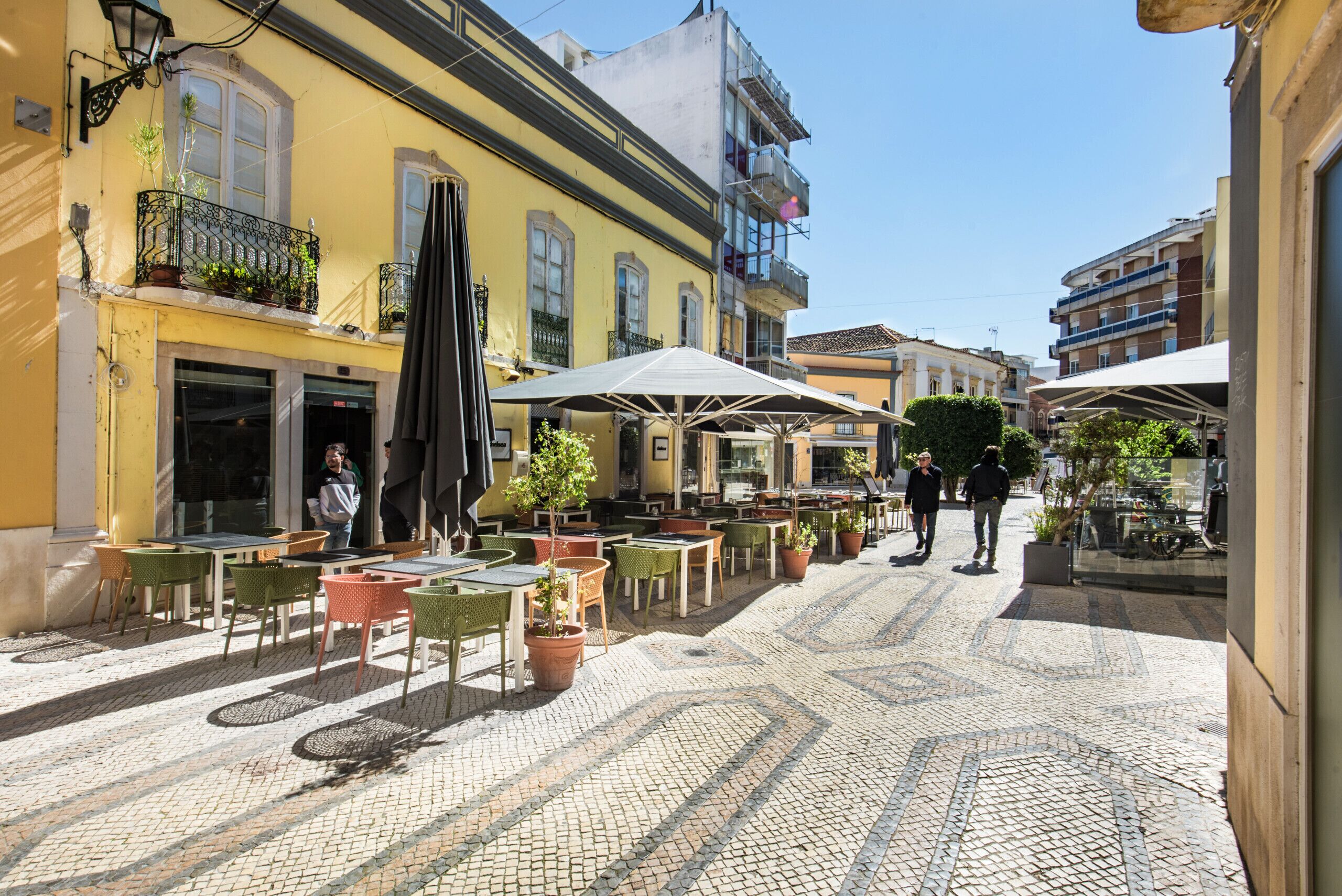 A cobblestone street with outdoor seating, potted plants, and a clear blue sky.