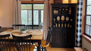 Un coin repas avec une table et des chaises en bois, un buffet noir contenant de l’ustensile de cuisine et une fenêtre avec des rideaux.