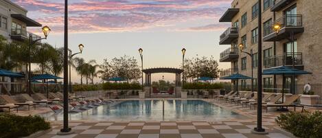 A pool area with lounge chairs and umbrellas, surrounded by buildings with balconies and a pergola.