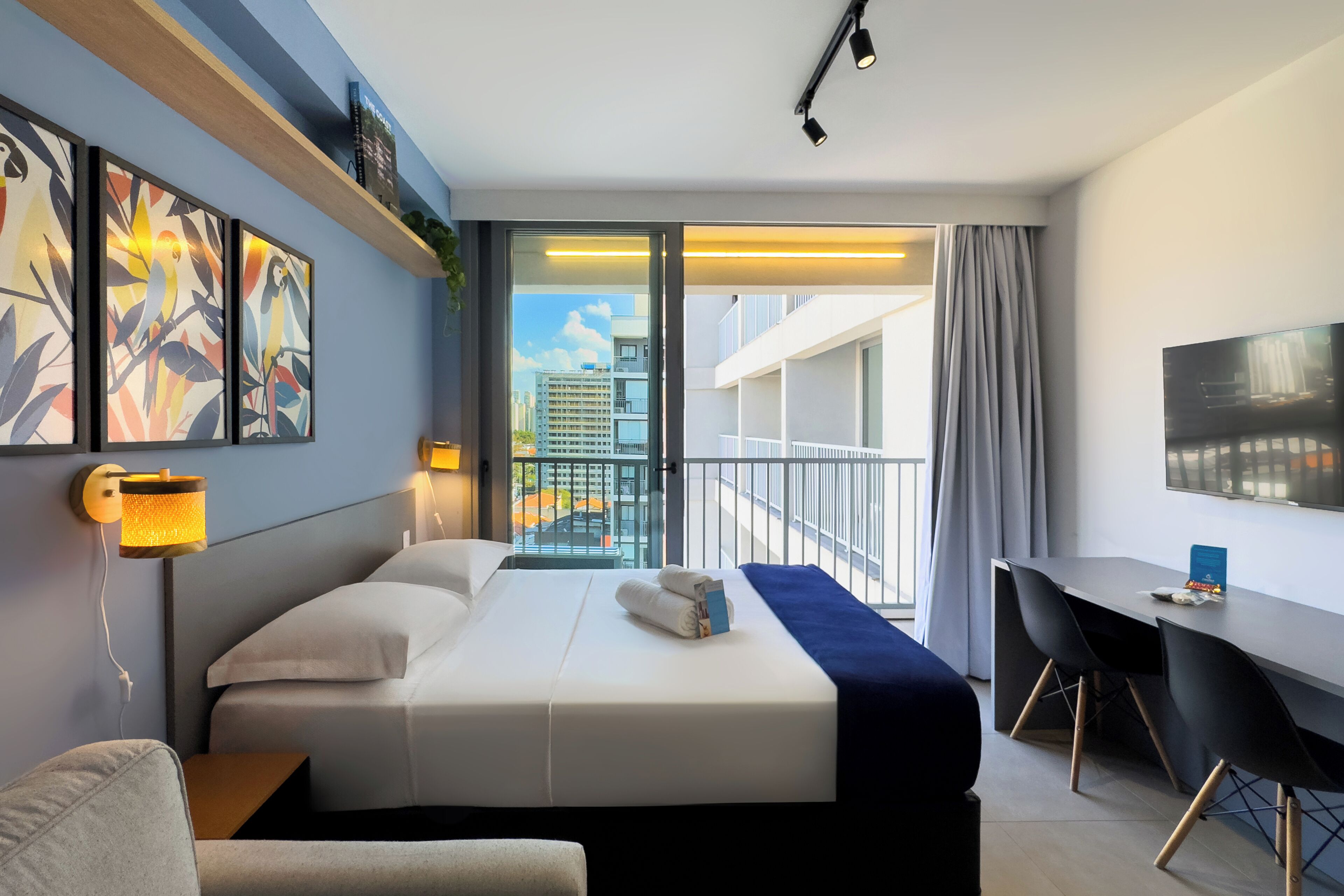 A modern hotel room with a bed, desk, and balcony with a city view.