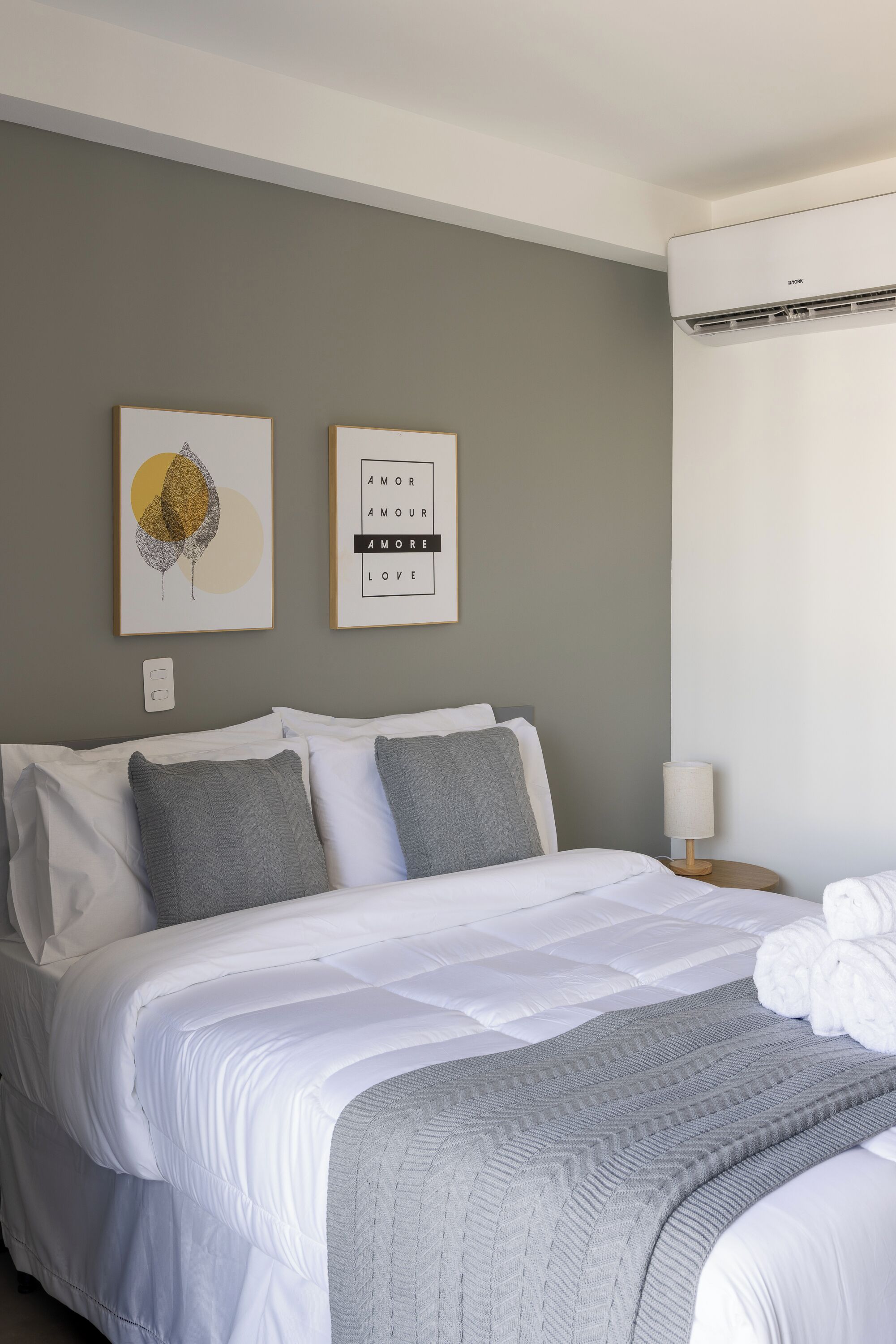 Een netjes opgemaakt bed met wit en grijs beddengoed, een grijze plaid en een luchtverkoeler aan de muur.