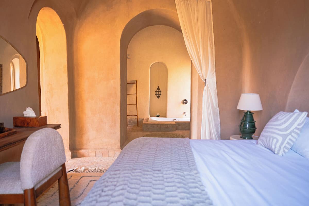 Un dormitorio con cama, escritorio, silla, lámpara y un baño visible a través de un arco.