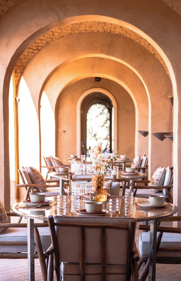 A dining area with a long table set for a meal, surrounded by arches and a view through a window. - Bliss Terra Janna (Marrakech)