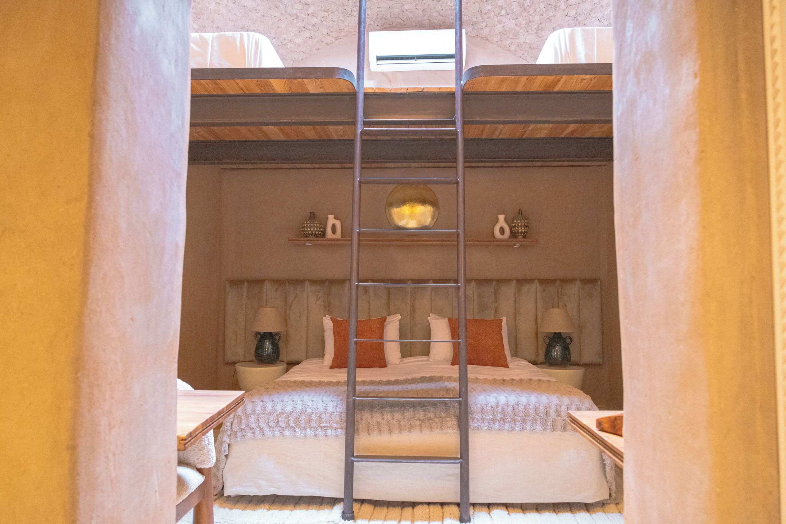 A bedroom with a bunk bed, a ladder, a bedside table, a lamp, and a wall-mounted shelf with decorative items.