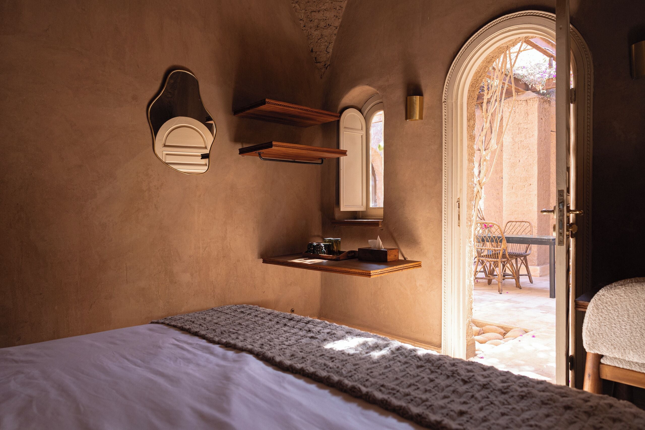 A bedroom with a bed, a wooden shelf with a mirror, and a door leading to an outdoor area with a table and chairs.
