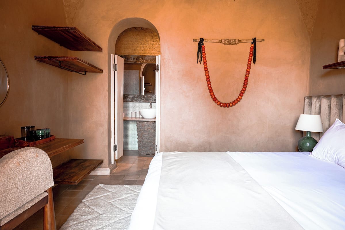 Un dormitorio con cama, un banco de madera, un collar de cuentas colgante y vistas a un baño a través de un umbral abierto.
