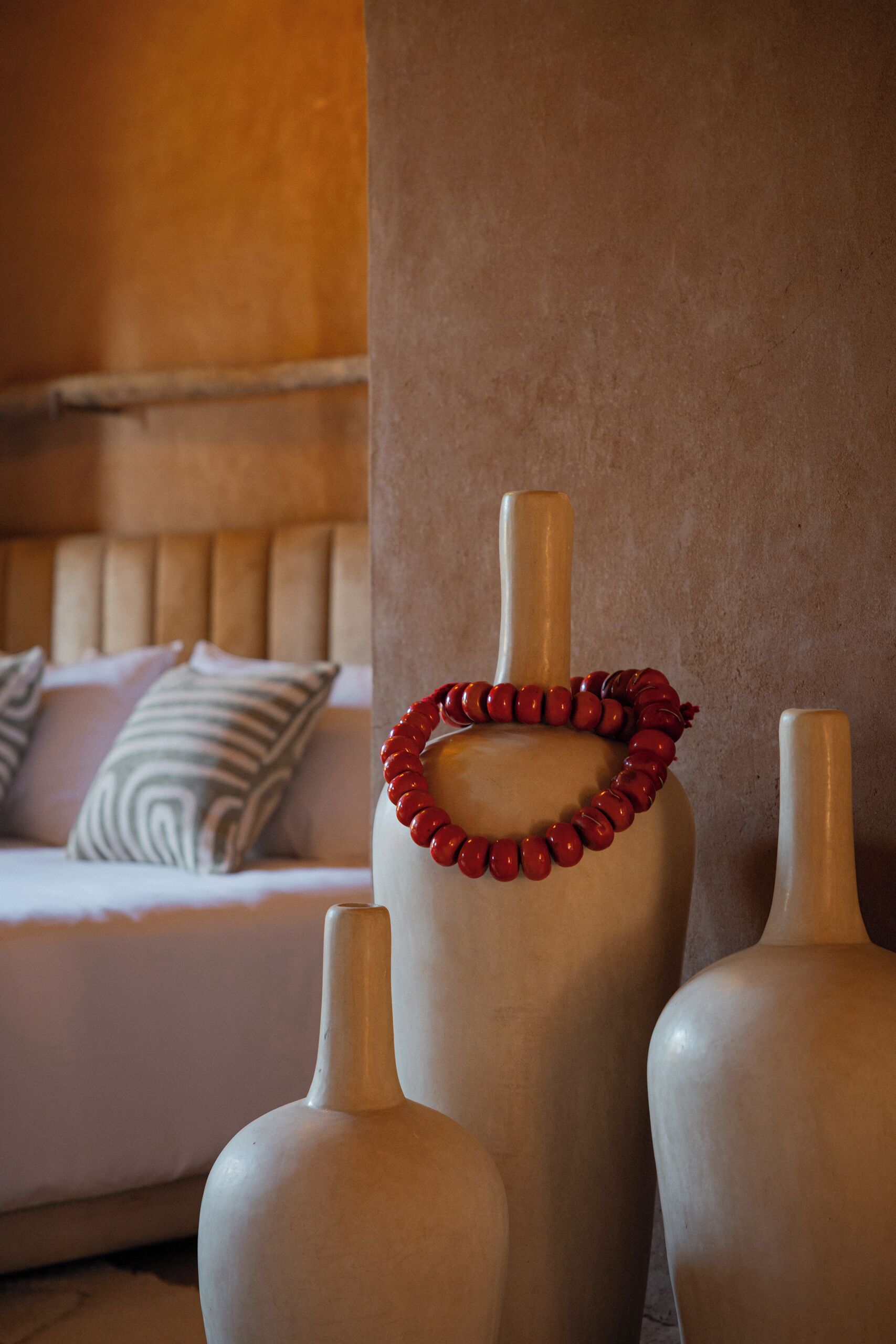 A room with a bed, pillows, and two vases with a red beaded necklace on top.