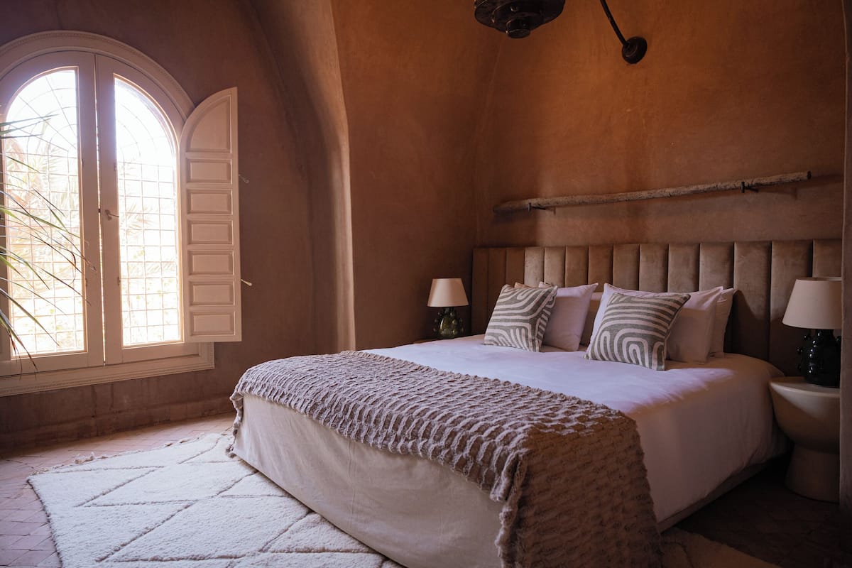 Un dormitorio con una cama grande, dos lámparas de noche, un cabecero y una ventana con contraventanas.
