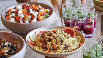Ein Tisch mit verschiedenen Gerichten, darunter ein Salat, Pasta und Spieße, sowie mehrere Schnapsgläser mit farbenfrohen Getränken.