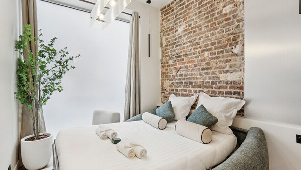 A modern hotel room with a brick accent wall, a large bed with white bedding, a potted plant, and a grey armchair. - Lavie Maison : Marais Luxury, Hoy Tub & A/C (Paris)