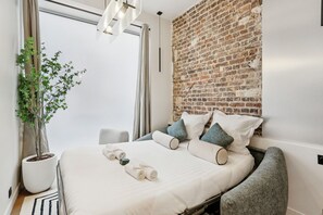 Et moderne hotelværelse med en murstensvæg, en stor seng med hvide sengetæger, en potteplante og en grå lænestol.