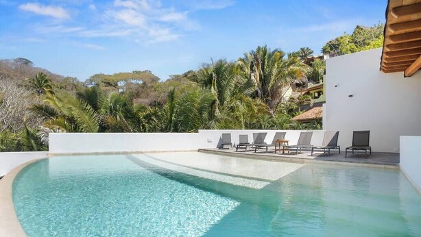 A clear blue swimming pool with lounge chairs and a white wall, surrounded by greenery and hills. - Northside Retreat - Infinity pool - Fast Wi-Fi (Sayulita)