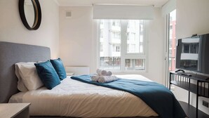Un letto rifatto con cuscini, un asciugamano arrotolato e una coperta blu. Una televisione su un mobile si trova sulla destra.