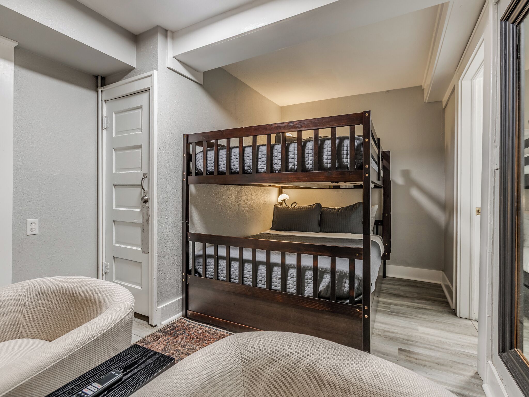 A bunk bed with a headboard, a bedside lamp, and a chair in a room with a white door and a small rug.